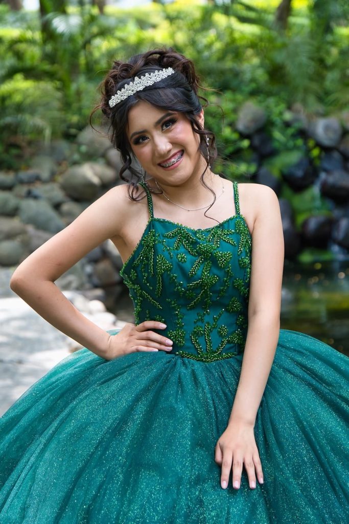 Quinceañera sonriendo en jardín natural con vestido verde brillante durante sesión fotográfica.